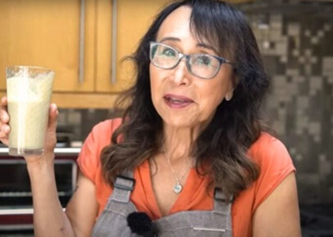 Woman with black hair and glasses holds up glass of protein shake as she stands in a kitchen.