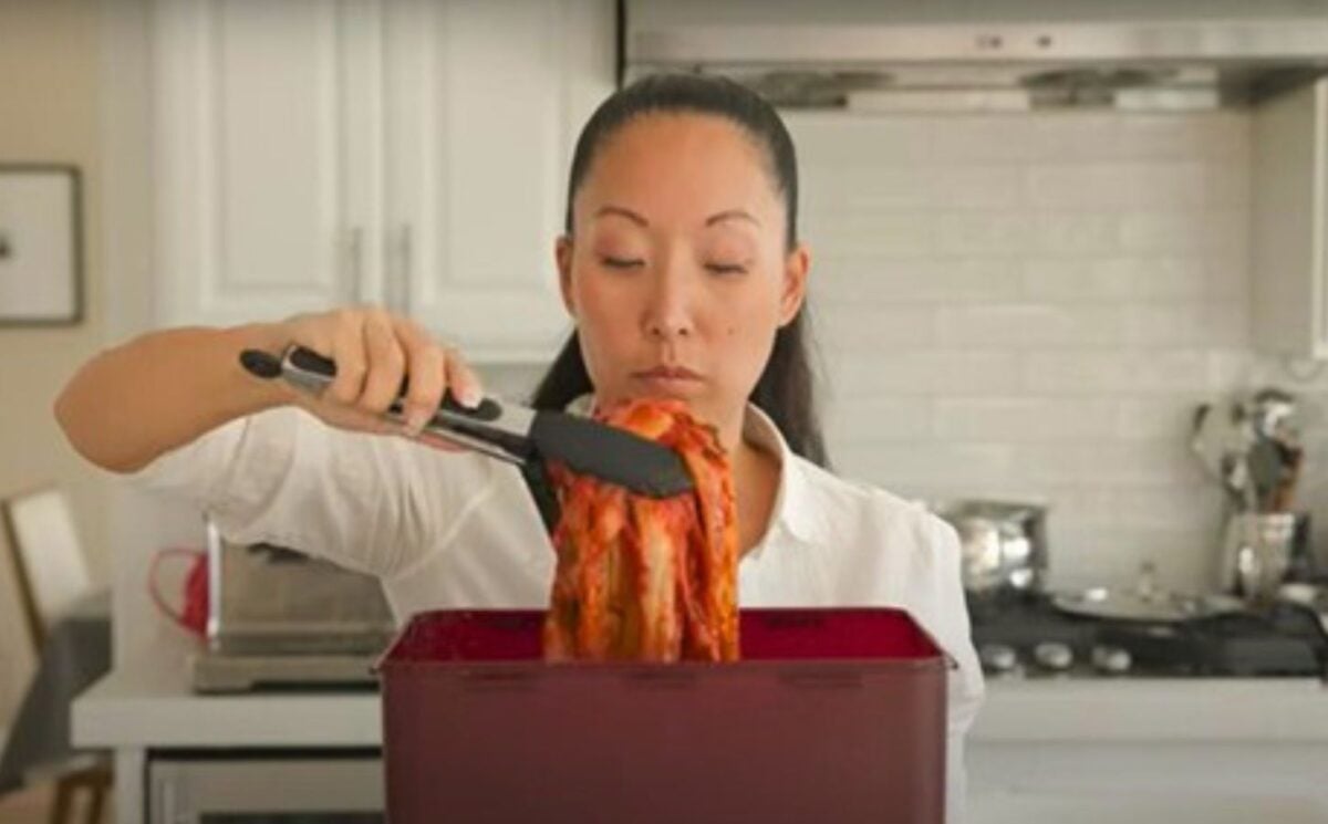 A woman in a white shirt lifts a large piece of kimchi with tongs from a red container in a kitchen.