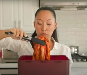 A woman in a white shirt lifts a large piece of kimchi with tongs from a red container in a kitchen.