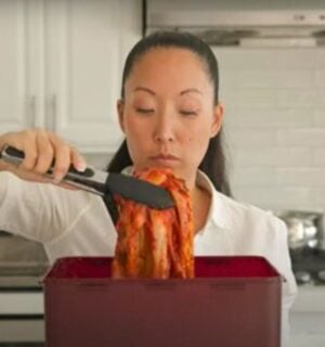 A woman in a white shirt lifts a large piece of kimchi with tongs from a red container in a kitchen.