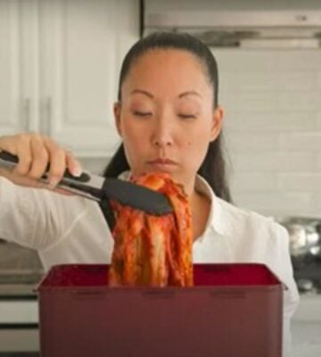 A woman in a white shirt lifts a large piece of kimchi with tongs from a red container in a kitchen.