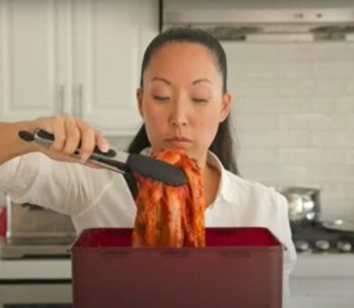 A woman in a white shirt lifts a large piece of kimchi with tongs from a red container in a kitchen.