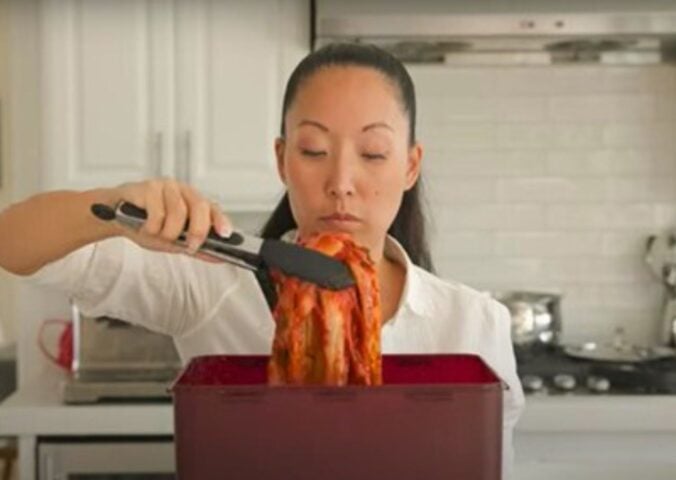 A woman in a white shirt lifts a large piece of kimchi with tongs from a red container in a kitchen.