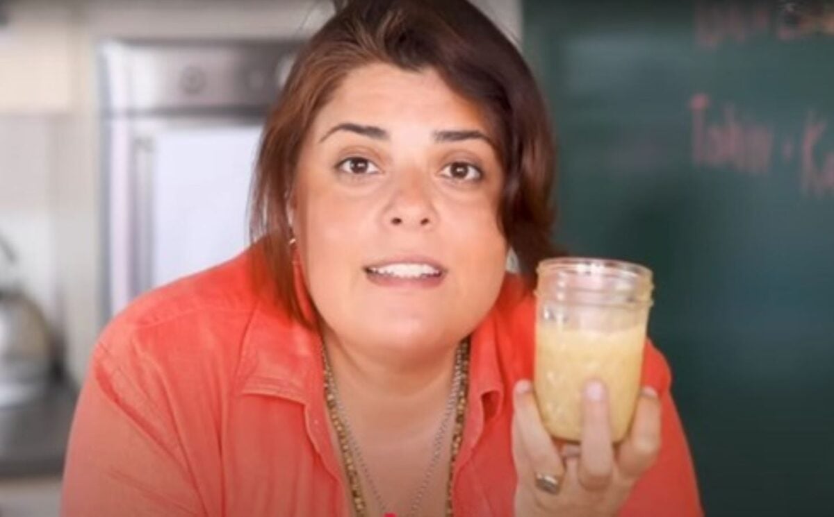 Brown-haired woman wearing orange shirt holds up glass jar of tahini as she stares intently into the camera.