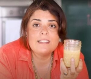 Brown-haired woman wearing orange shirt holds up glass jar of tahini as she stares intently into the camera.