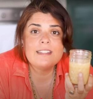 Brown-haired woman wearing orange shirt holds up glass jar of tahini as she stares intently into the camera.