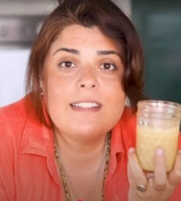 Brown-haired woman wearing orange shirt holds up glass jar of tahini as she stares intently into the camera.