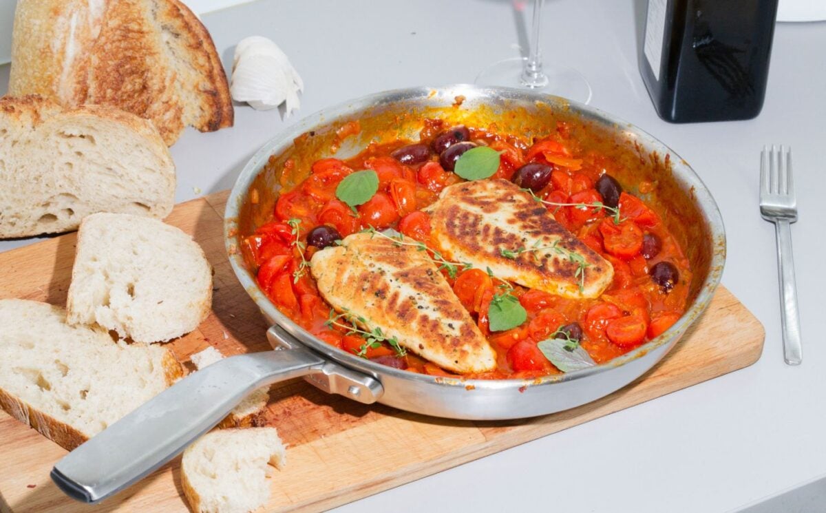A steel pan containing two plant-based cod filets in tomato sauce sits on top of a wooden cutting board next to a loaf of bread with a few slices cut off.