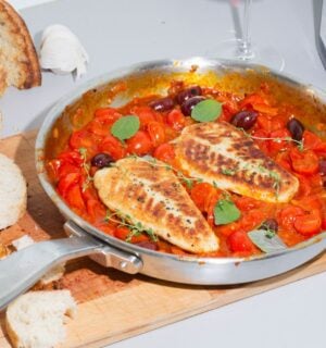 A steel pan containing two plant-based cod filets in tomato sauce sits on top of a wooden cutting board next to a loaf of bread with a few slices cut off.