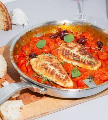 A steel pan containing two plant-based cod filets in tomato sauce sits on top of a wooden cutting board next to a loaf of bread with a few slices cut off.