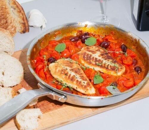 A steel pan containing two plant-based cod filets in tomato sauce sits on top of a wooden cutting board next to a loaf of bread with a few slices cut off.