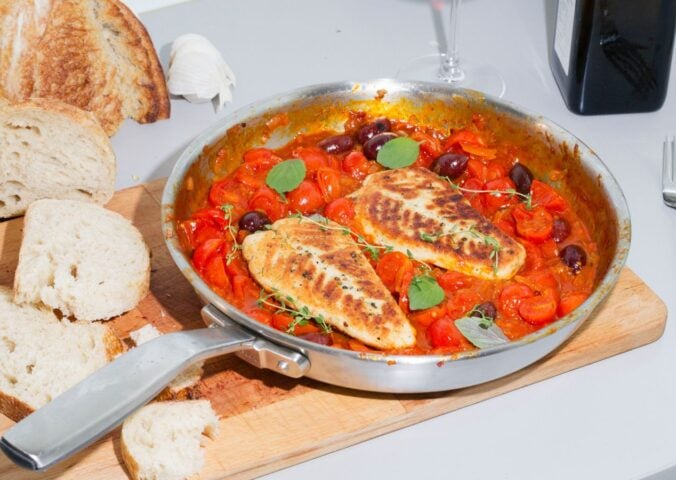 A steel pan containing two plant-based cod filets in tomato sauce sits on top of a wooden cutting board next to a loaf of bread with a few slices cut off.