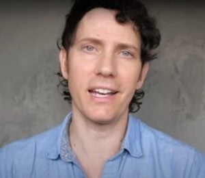 Wearing a light blue shirt over a grey background, Mic the Vegan speaks about plant-based protein.