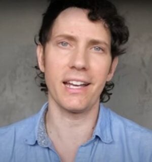 Wearing a light blue shirt over a grey background, Mic the Vegan speaks about plant-based protein.