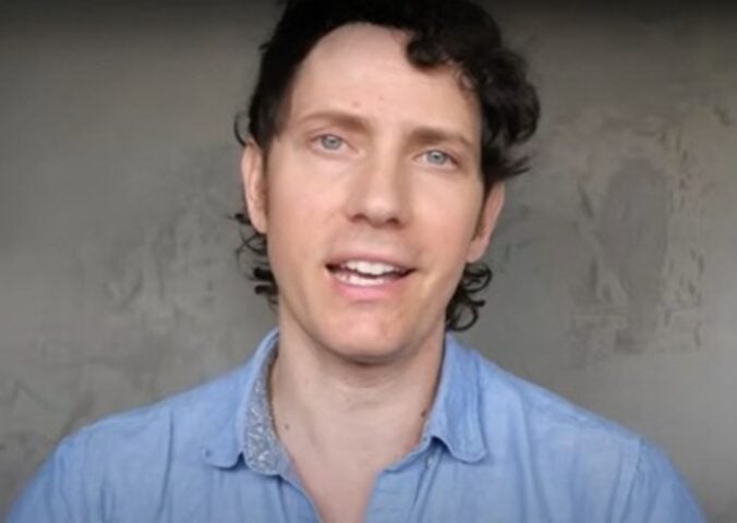 Wearing a light blue shirt over a grey background, Mic the Vegan speaks about plant-based protein.