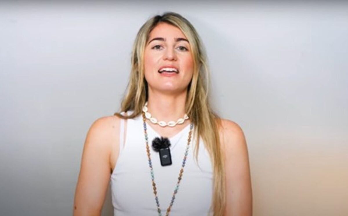 Charlotte, from Vegan Explores, is wearing a white sleeveless top and standing before a greyish background while sharing what she has learned about traveling as a vegan