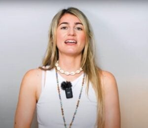 Charlotte, from Vegan Explores, is wearing a white sleeveless top and standing before a greyish background while sharing what she has learned about traveling as a vegan