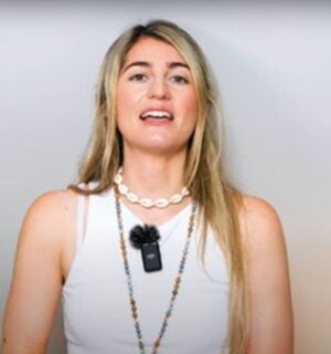 Charlotte, from Vegan Explores, is wearing a white sleeveless top and standing before a greyish background while sharing what she has learned about traveling as a vegan