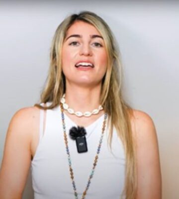 Charlotte, from Vegan Explores, is wearing a white sleeveless top and standing before a greyish background while sharing what she has learned about traveling as a vegan
