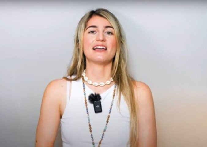 Charlotte, from Vegan Explores, is wearing a white sleeveless top and standing before a greyish background while sharing what she has learned about traveling as a vegan