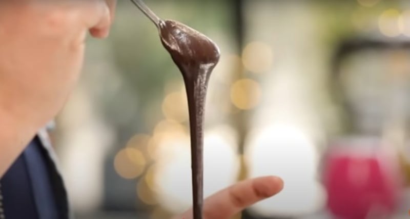 Hand holds up spoon dripping with a chocolatey, Nutella-like paste over a blurred background.