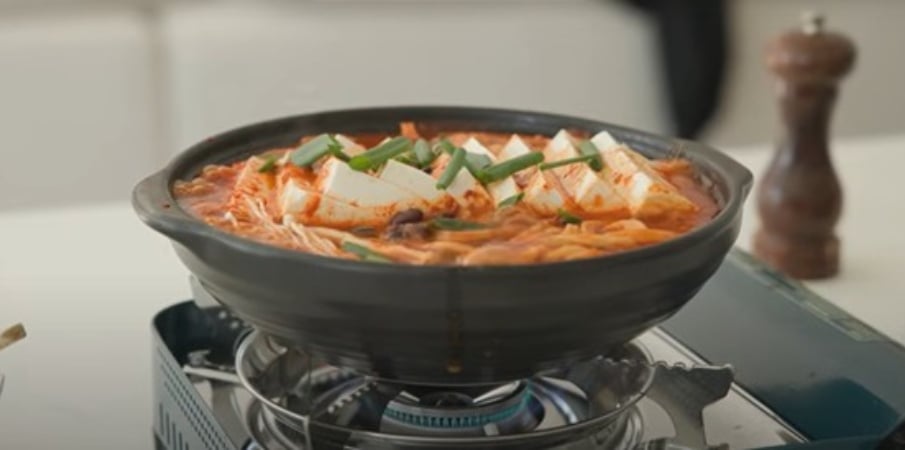 A bubbling pot of kimchi jjigae simmers on a portable gas stove, topped with sliced tofu and chopped scallions.