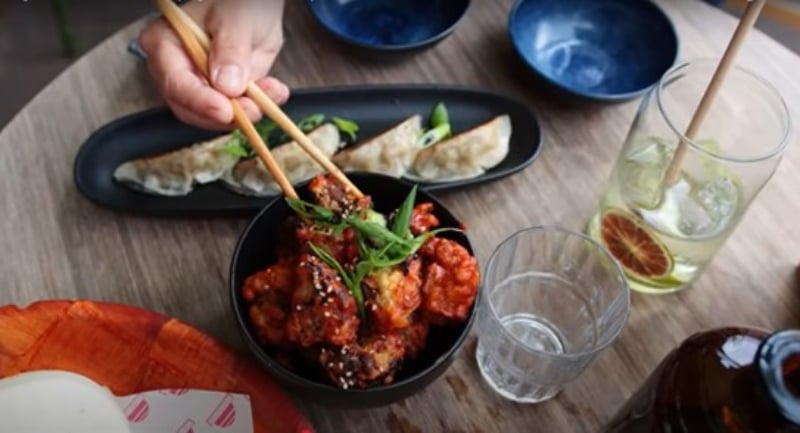 A hand uses chopsticks to pick up a cauliflower wing from a bowl. Behind the wings is a black plate with four gyozas, and there are two glasses on the table and several other little plates and a black bottle