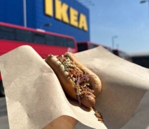Photo shows someone holding up a vegan IKEA hotdog topped with La Vie bacon bits, in front of an IKEA store