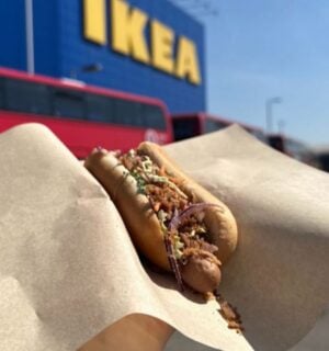 Photo shows someone holding up a vegan IKEA hotdog topped with La Vie bacon bits, in front of an IKEA store