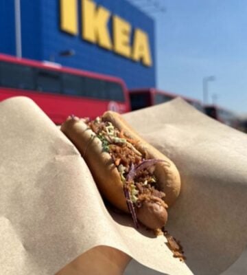 Photo shows someone holding up a vegan IKEA hotdog topped with La Vie bacon bits, in front of an IKEA store