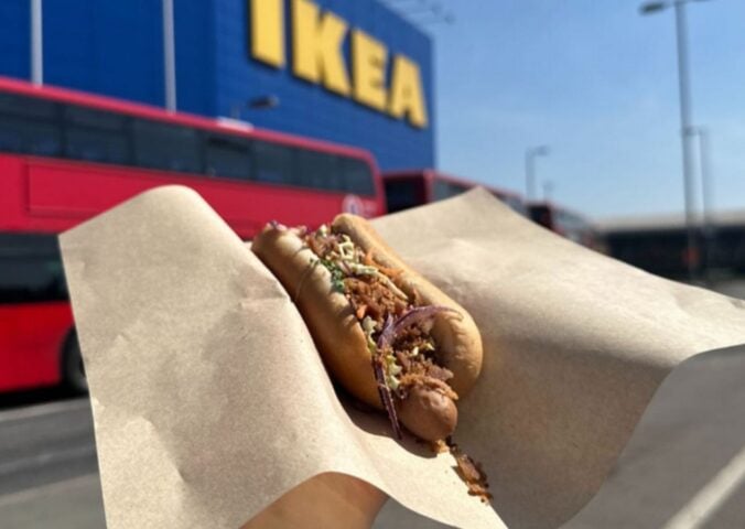 Photo shows someone holding up a vegan IKEA hotdog topped with La Vie bacon bits, in front of an IKEA store