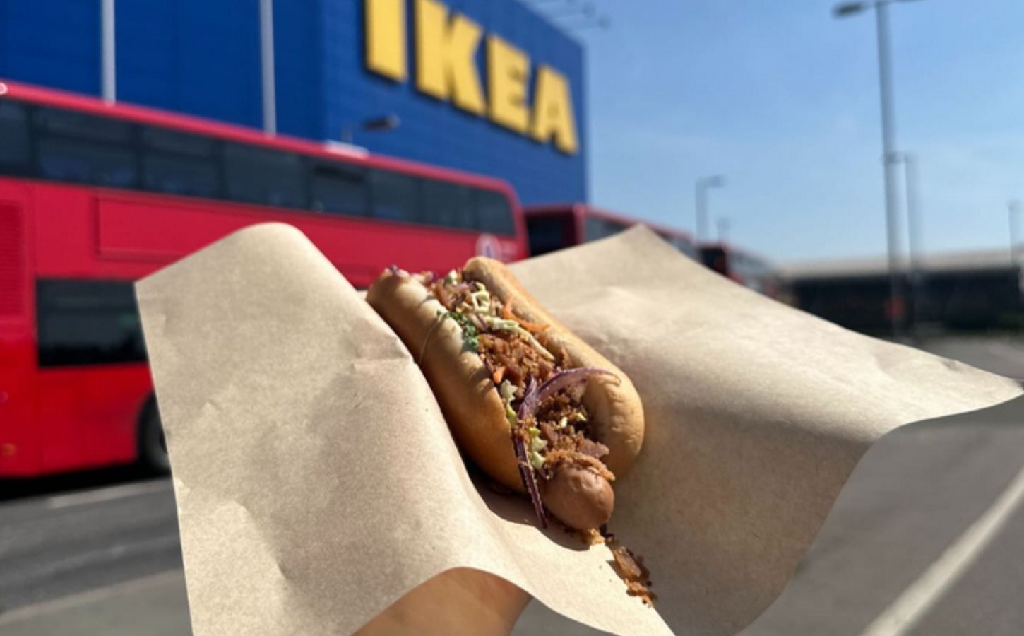 Photo shows someone holding up a vegan IKEA hotdog topped with La Vie bacon bits, in front of an IKEA store