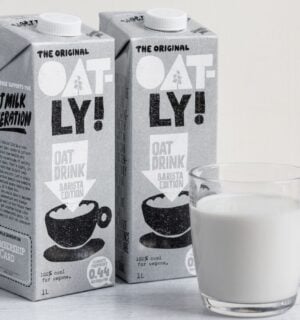 Photo shows two cartons of Oatly Barista next to a glass mug of oat milk