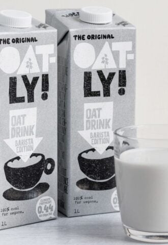 Photo shows two cartons of Oatly Barista next to a glass mug of oat milk