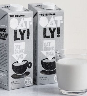 Photo shows two cartons of Oatly Barista next to a glass mug of oat milk