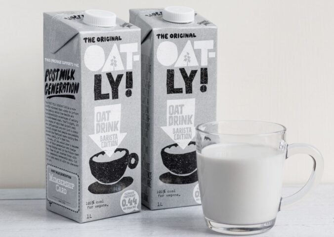Photo shows two cartons of Oatly Barista next to a glass mug of oat milk