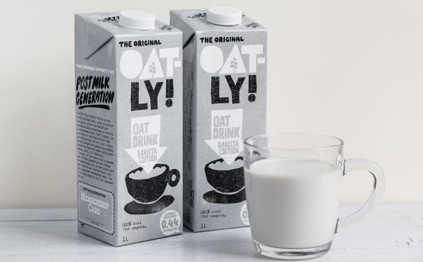 Photo shows two cartons of Oatly Barista next to a glass mug of oat milk