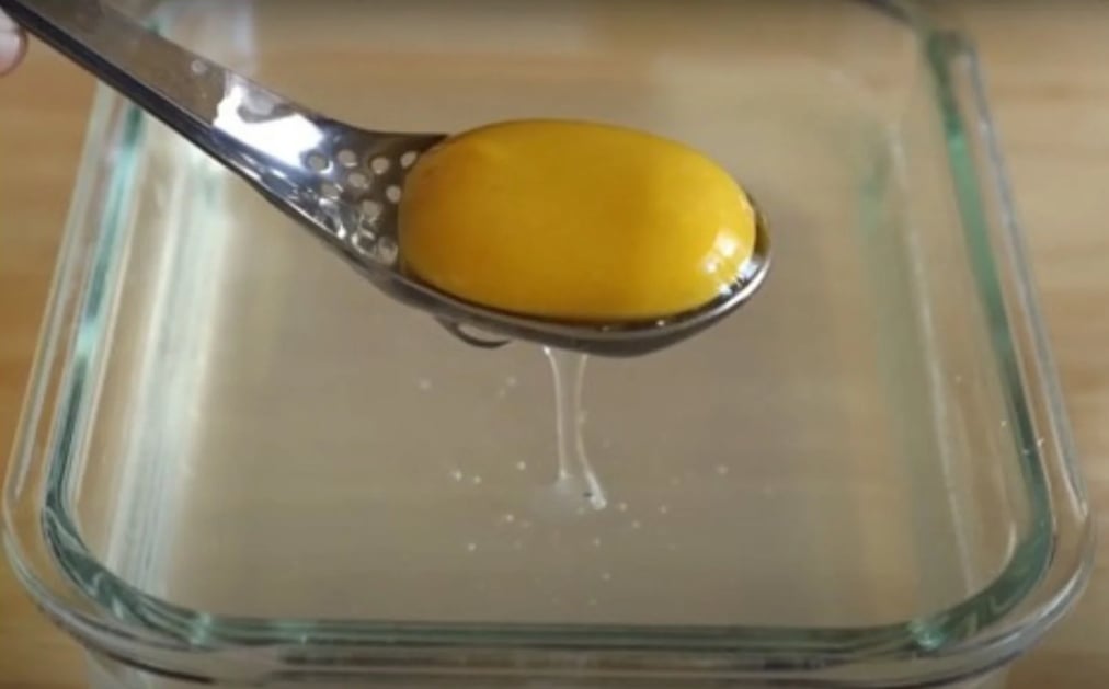 A skimmer spoon lifts a plant-based egg yolk out of the transparent, liquid mixture in a glass baking dish.
