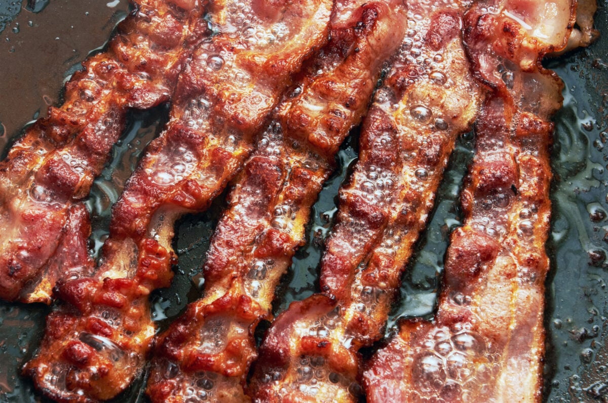 Photo shows a pan of streaky bacon cooking in fat and oil