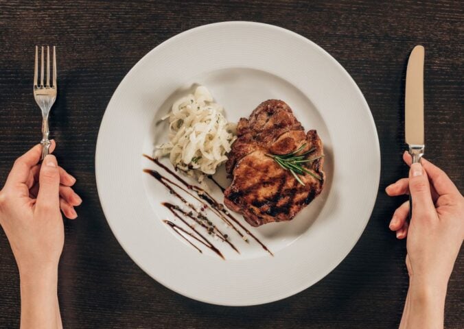 Photo shows a woman's hands holding a knife and fork next to a plate of steak and mashed potatoes
