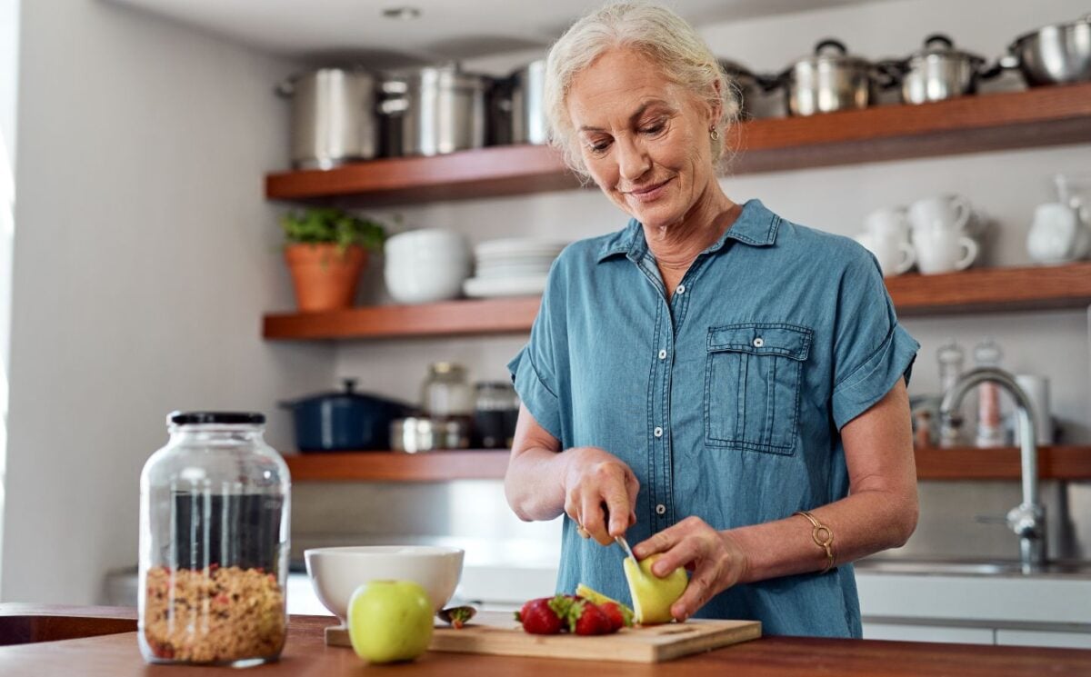 Photo shows an older woman chopping fresh fruit and vegetables in a kitchen