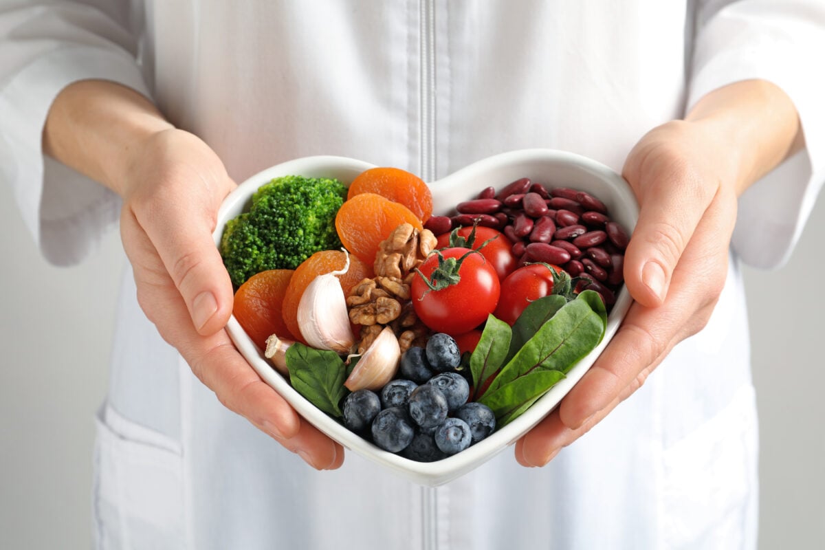 Photo shows someone in a white coat holding a heart-shaped bowl full of nutritious plant-based foods, including blueberries, garlic, greens, beans, and tomatoes