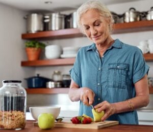 Photo shows an older woman chopping fresh fruit and vegetables in a kitchen