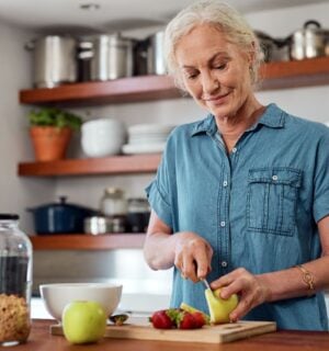 Photo shows an older woman chopping fresh fruit and vegetables in a kitchen