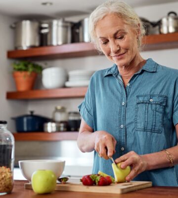 Photo shows an older woman chopping fresh fruit and vegetables in a kitchen