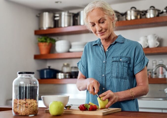 Photo shows an older woman chopping fresh fruit and vegetables in a kitchen