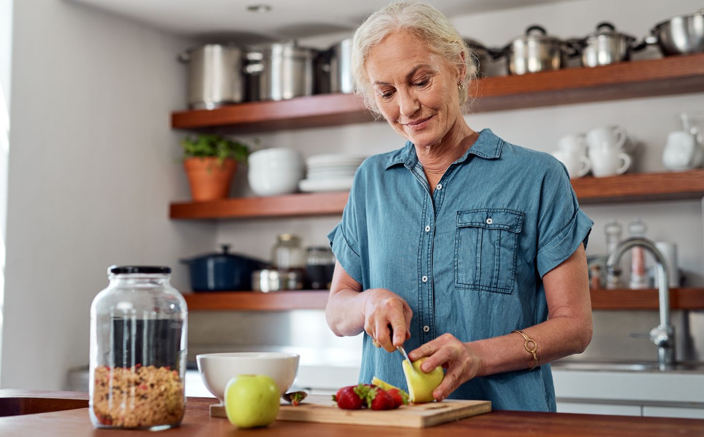 Photo shows an older woman chopping fresh fruit and vegetables in a kitchen