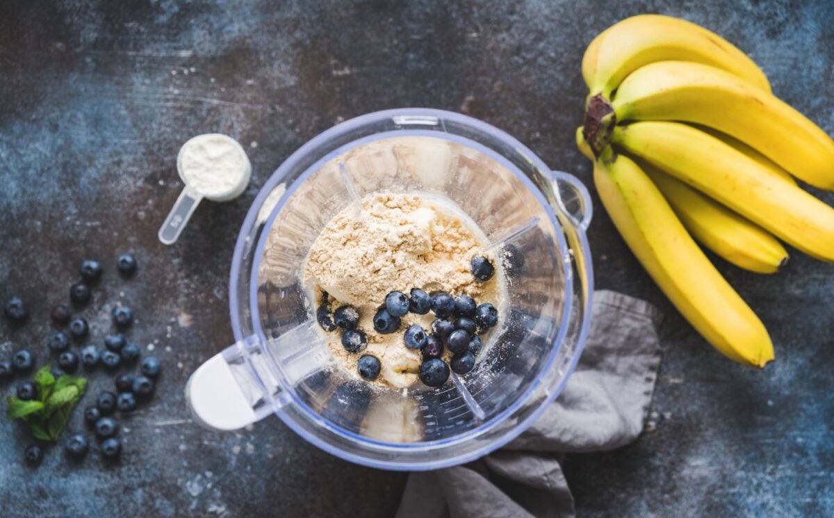 Photo shows an open blender from above containing blueberries and vegan protein powder next to a bunch of a bananas