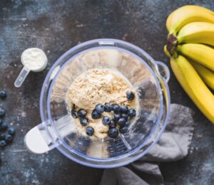 Photo shows an open blender from above containing blueberries and vegan protein powder next to a bunch of a bananas