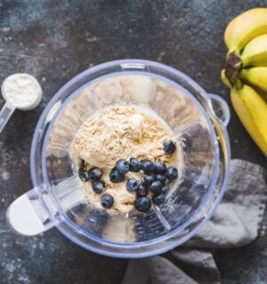 Photo shows an open blender from above containing blueberries and vegan protein powder next to a bunch of a bananas